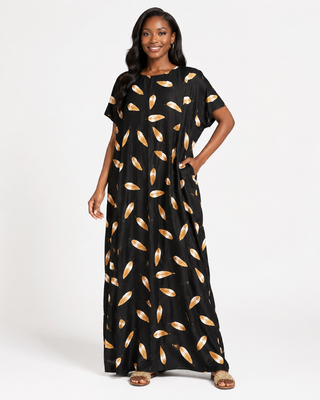 The image features a woman wearing a long, flowing maxi dress in black with an eye-catching pattern of gold and orange leaf shapes. The dress has short sleeves and a relaxed fit, giving it a comfortable yet stylish appearance. The woman is posing confidently with one hand in her pocket, showcasing the dress's design.
