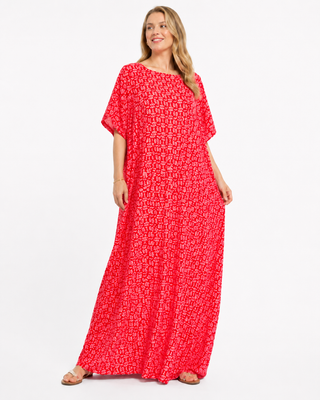 The image shows a woman wearing a long, flowing red dress with a patterned design. The dress has short sleeves and features a loose fit, reaching down to the floor. The background is plain and light-colored, emphasizing the vibrant color of the dress.
