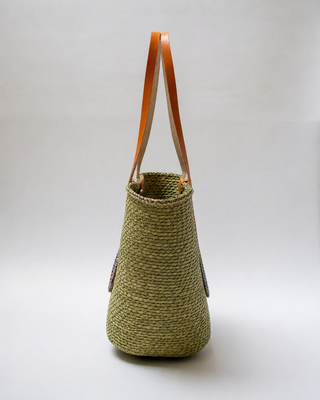 Woven Straw Kiondoo basket with two orange leather handles and a circular decorative patch featuring colorful beads on the front