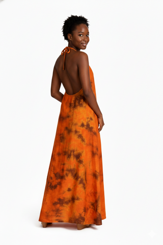 A model is wearing a long, flowy dress in shades of orange with a tie-dye pattern. The dress features a halter neck design and flows to the floor, showcasing a relaxed and elegant style. The model stands confidently, highlighting the dress's vibrant colors and lightweight fabric.
