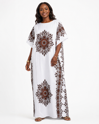 The image features a woman wearing a long, white kaftan dress with intricate brown and red patterns on the sleeves and sides. The dress has a loose fit and falls to the ankles, providing a comfortable and stylish appearance. The woman is standing confidently with a slight smile.